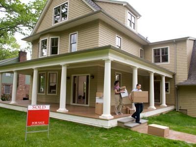 Couple carrying boxes out of a house with Sold sign on lawn